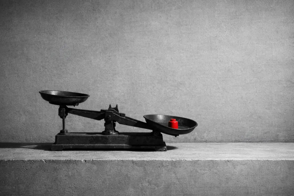 An old two-pan balance scale on a concrete ledge, tipped by a single red weight in the lower pan